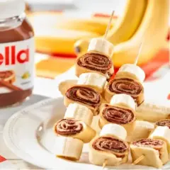 Naleśniki z nutellą i bananem na patyczkach, obok słoik kremu. Idealna przekąska dla każdego.